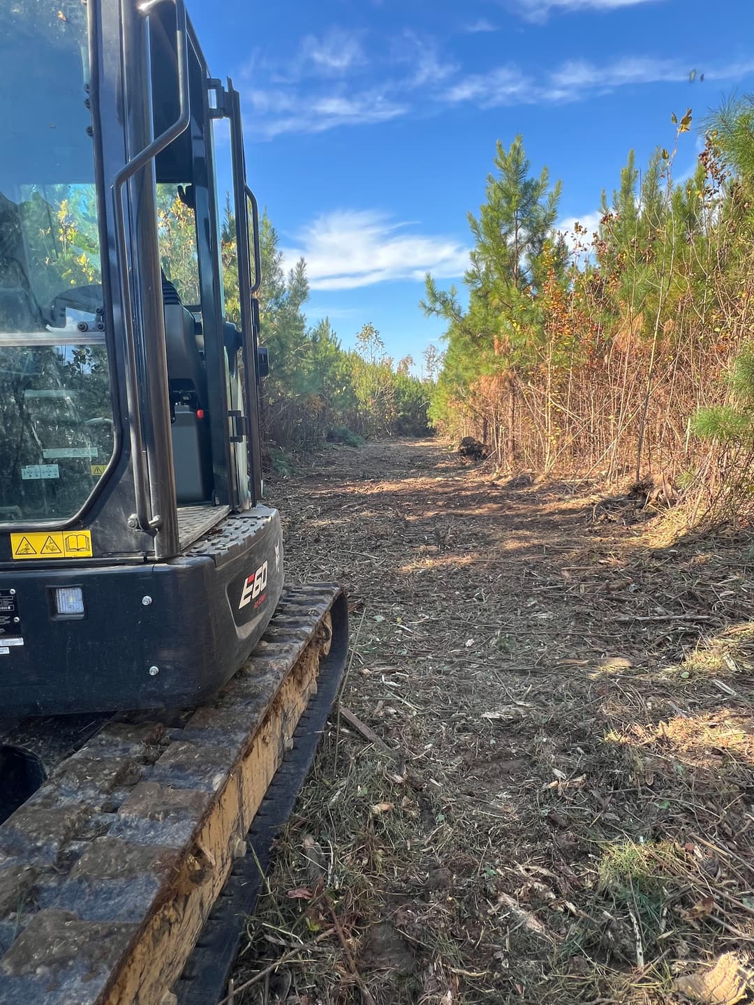 Excavator clearing a wooded path under a blue sky with green foliage surrounding the trail.