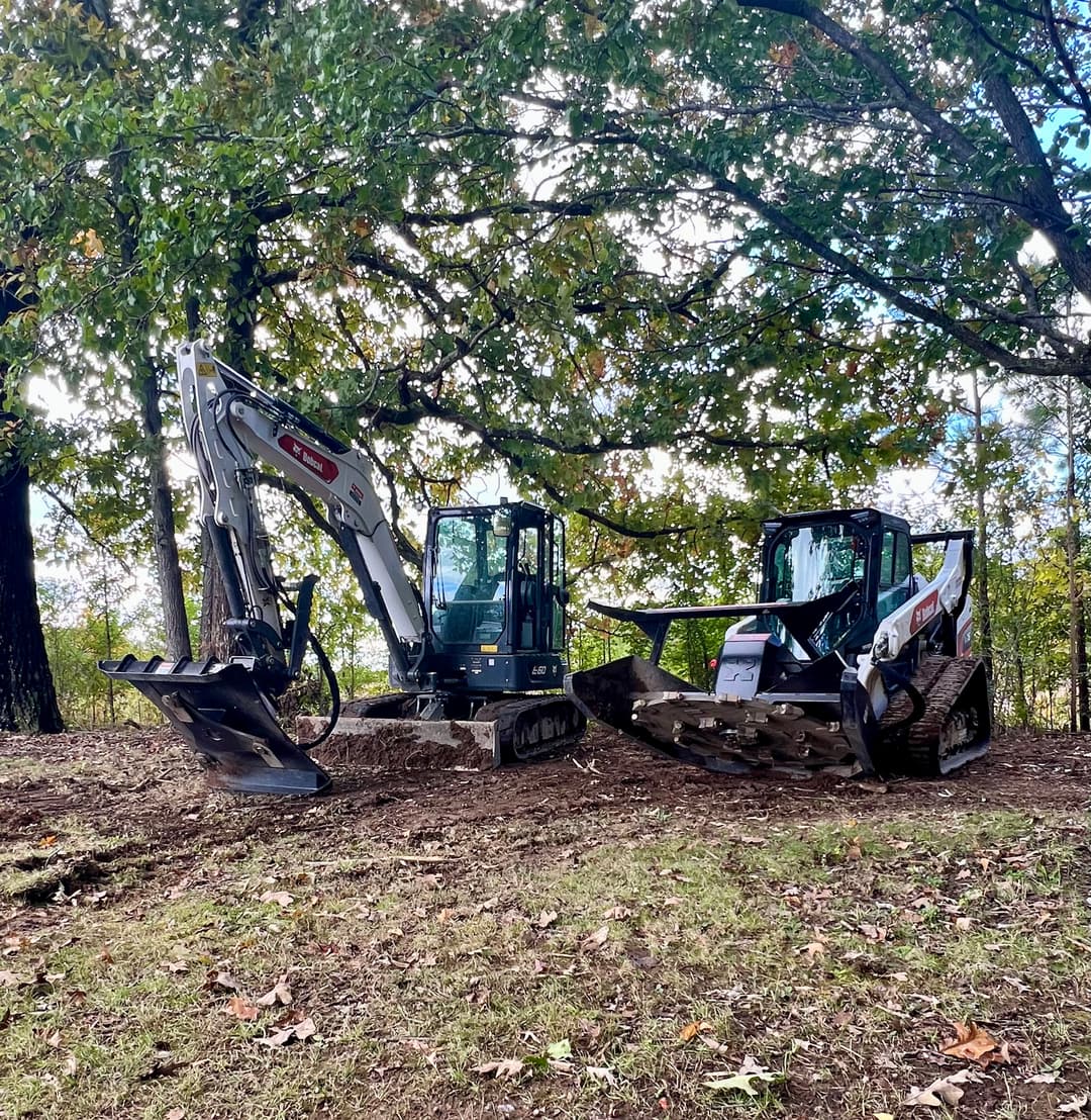Excavators positioned on a wooded site, surrounded by autumn foliage and trees.