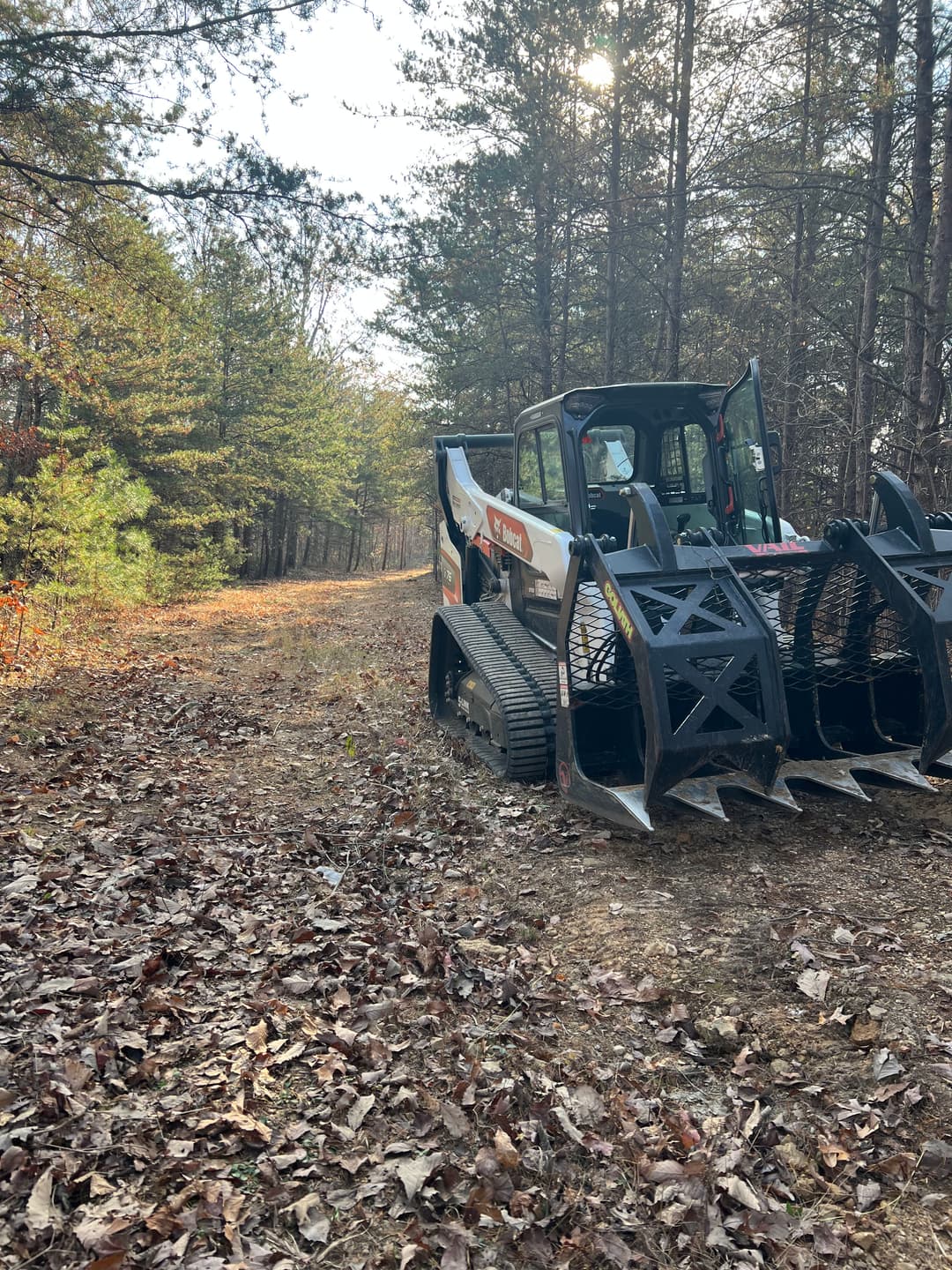 Bobcat skid steer clearing a woodland path with fallen leaves in autumn scenery.