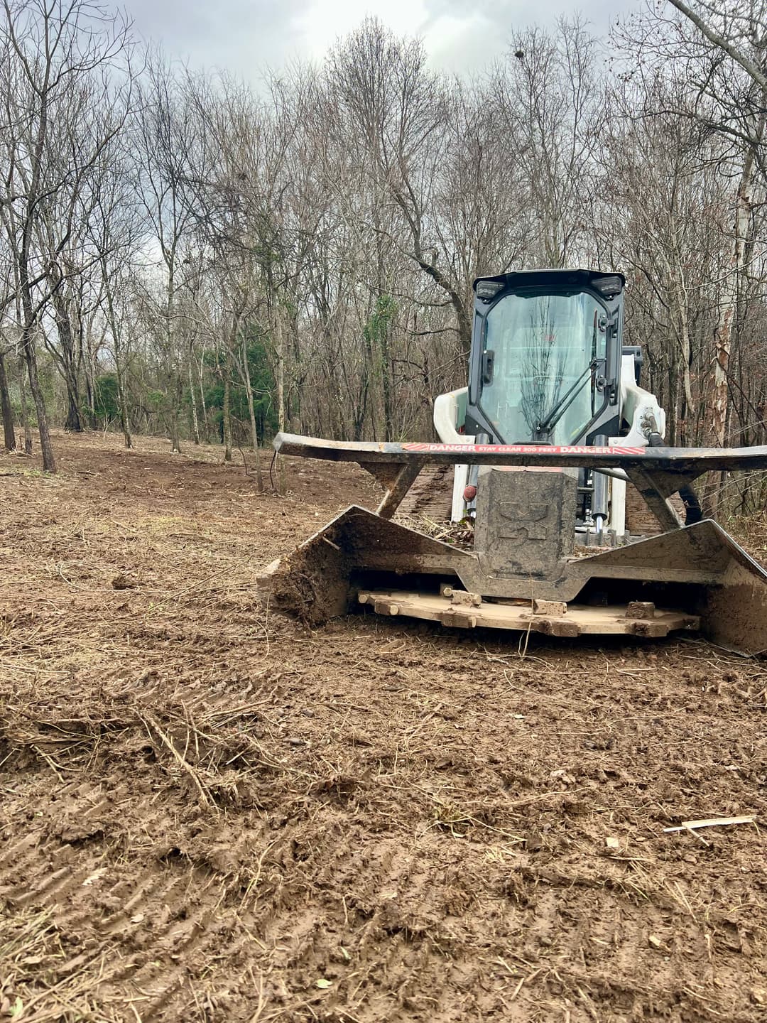 Bobcat skid steer with brush cutter clears land in a wooded area, showing freshly disturbed soil.